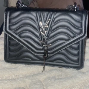 Svart axelväska YSL - Snygg svart axelväska från YSL med quiltat mönster och silverdetaljer. Väskan har en kedjerem och ett elegant YSL-emblem på framsidan. Jättefin väska som tyvärr inte kommit till användning 🌸