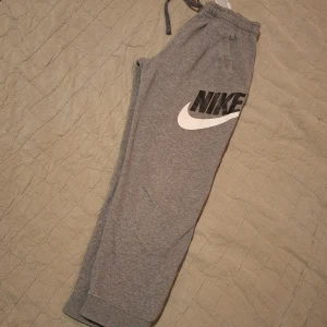 Grå mjukisbyxor från Nike - Säljer ett par grå mjukisbyxor från Nike med elastisk midja och snörning. Byxorna har en stor Nike-logga på benet och är perfekta för en avslappnad stil. De är tillverkade i mjukt material för extra komfort.