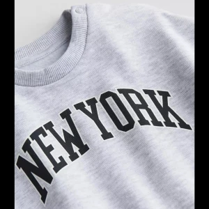 Grå sweatshirt med New York-tryck - Snygg grå sweatshirt med svart New York-tryck på bröstet. Tröjan har en rund halsringning och långa ärmar, perfekt för en avslappnad stil. Passar bra till jeans eller leggings för en cool vardagslook.