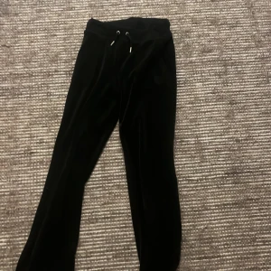 Svarta mjukisbyxor från Juicy Couture - Snygga svarta mjukisbyxor från Juicy Couture i mjukt material. Byxorna har en bekväm passform med snörning i midjan och stilrena fickor med knappar bak. Perfekta för en avslappnad look.