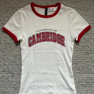 Vit t-shirt med Cambridge-tryck  - Snygg vit t-shirt från Divided med rött Cambridge-tryck på bröstet. Den har röda kanter vid hals och ärmslut. Ger en snygg 90-tals vibe!! 🎸💖 Perfekt för sommardagar! Använd max 3 gånger så är i helt nytt skick. Skriv gärna till mig vid intresse 😊