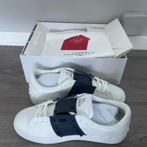 Valentino Garavani Opens - Snygga vita sneakers från Valentino Garavani med navy strecket över snörningen. Skorna har en stilren design med ikoniska nitar på remmen och en bekväm sula. Perfekta för att ge en lyxig touch till din outfit. Fler bilder kan skickas privat. Nytt skick.