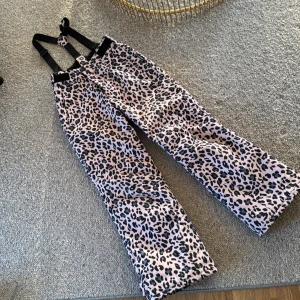 Leopardmönstrade skidbyxor med hängslen - Snygga och trendiga täckbyxor med leopardmönster i rosa och svart. Byxorna har justerbara svarta hängslen och man kan justera i midjan och runt fötterna med kardborreband. De är mycket sparsamt användna och i gott skick. En liten tråd har släppt på insidan vid höger fot. Lätt att åtgärda, påverkar ej funktionen.