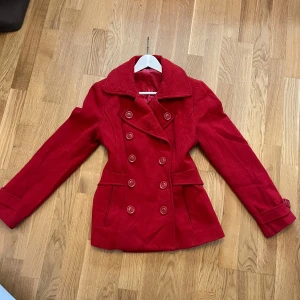 Röd kappa med knappar - Snygg röd kappa med dubbelknäppning och bred krage. Jackan har långa ärmar och en figursydd passform som ger en stilren look. Perfekt för kyligare dagar.