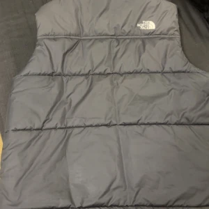 Svart dunväst från The North Face storlek L - Säljer en svart dunväst från The North Face med dragkedja framtill. Västen har en quiltad design och är perfekt för kyligare dagar. Märkesloggan är synlig på bröstet och ryggen. St L