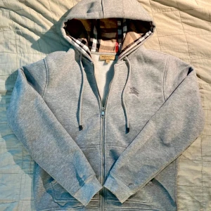 Grå Burberry hoodie - Säljer en grå hoodie från Burberry med dragkedja och huva. Insidan av huvan har det klassiska rutiga fodret. Tröjan har långa ärmar och snören vid huvan. Skön och stilren för vardagen.