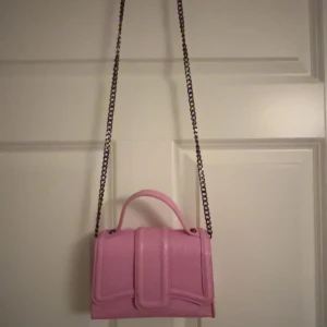 Rosa axelväska med kedja - Snygg rosa axelväska med en stilren kedjerem och ett praktiskt handtag. Väskan har en elegant design med en strukturerad yta och en smidig stängning framtill. Perfekt för att ge din outfit en färgklick!