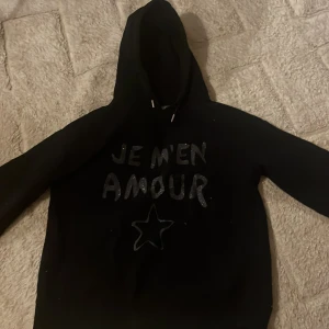 Svart hoodie från Gina Tricot - Snygg svart hoodie från Gina Tricot med texten 'JE M'EN AMOUR' i glittriga bokstäver på framsidan. Tröjan har en klassisk huva och långa ärmar, perfekt för en avslappnad stil.