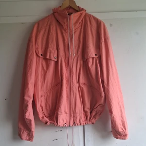 Rosa vindjacka med dragkedja - Säljer en snygg korall vindjacka med dragkedja framtill. Jackan har en huva med snörning och två stora fickor med knappar på bröstet. Den har en avslappnad passform och elastisk nederkant. oversize passar de flesta. äkta vintage.