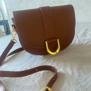 Brun axelväska med gulddetaljer - Väska i "saddle bag" stil i klassisk brun färg. Den har en innerficka med dragkedja och ett annat litet fack. En längre axelrem med justerbar längd och en kortare tillhör så det går bra att ha väskan som cross body eller bara som en shoulder bag. Väldigt fin väska, helt oanvänd!  Mått: 22x8,5 x17cm
