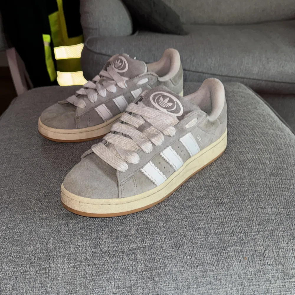 Säljer ett par gråa Adidas sneakers i storlek 43. Skorna är i mocka med vita detaljer och snörning. De har en gummisula som är lite sliten men fortfarande i bra skick. Perfekta för vardagsbruk! Tillkommer extra snören, tyvärr finns ej box eller kvitto.. Skor.