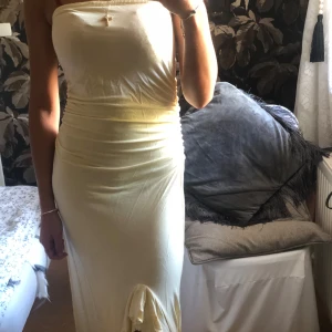 Champagnefärgad axelbandslös långklänning - Säljer en elegant champagnefärgad långklänning som är axelbandslös och har en snygg slits framtill. Perfekt för festliga tillfällen! Klänningen har en tight passform som framhäver figuren.