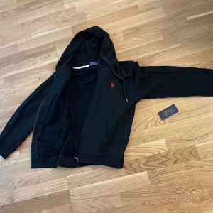 Svart hoodie från Ralph Lauren - Säljer en snygg och stilren svart hoodie från Ralph Lauren. Den har en dragkedja framtill och en klassisk röd logga på bröstet. Perfekt för både höst och vinter, och den är superbekväm att ha på sig. Passar till alla tillfällen! kostade mig mycket men säljer för så billigt för de inte passar längre mig har använt den i ungefär 4 månader 