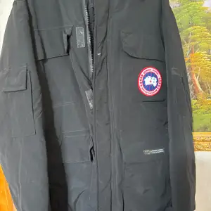 Säljer en svart vinterjacka från Canada Goose. Den är perfekt för kalla dagar med sitt tjocka och varma material. Jackan har en dragkedja och flera praktiska fickor. Märkets ikoniska logga syns tydligt på bröstet. Passar perfekt för vinterns alla äventyr!