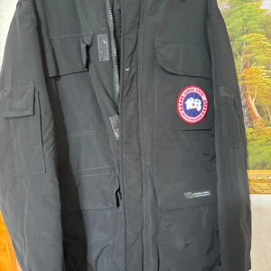 Svart vinterjacka från Canada Goose - Säljer en svart vinterjacka från Canada Goose. Den är perfekt för kalla dagar med sitt tjocka och varma material. Jackan har en dragkedja och flera praktiska fickor. Märkets ikoniska logga syns tydligt på bröstet. Passar perfekt för vinterns alla äventyr!