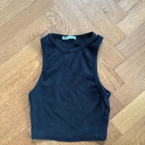 Svart ribbad linnetopp från Zara - Säljer en snygg svart ribbad linnetopp från Zara. Den är ärmlös och croppad, perfekt för sommardagar eller som ett lager under en jacka. Toppen är i ett mjukt och stretchigt material som sitter skönt mot kroppen.