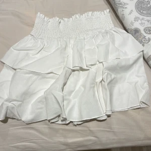 Vit volangkjol - Säljer en superfin vit kjol med volanger. Den har en elastisk midja som gör den bekväm och lätt att bära. Perfekt för sommardagar eller en festlig kväll. Kjolen är i bra skick och redo för nya äventyr!