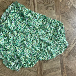 Blommig grön kjol - Säljer en superfin blommig kjol i grönt med inslag av lila och vitt. Kjolen har en härlig volangdetalj och är perfekt för sommarens alla fester. Materialet känns lätt och luftigt, vilket gör den idealisk för varma dagar. Passar perfekt för en dag på stan eller en picknick i parken!