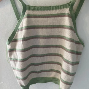 Randig stickad linne i grönt och vitt - Säljer en superfin stickad linne med gröna och vita ränder. Den har en enkel och stilren design med gröna kanter och axelband. Perfekt för sommardagar eller som ett lager under en jacka.