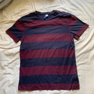 Randig t-shirt från H&M Divided - Säljer en snygg randig t-shirt från H&M Divided i storlek M. Den har breda ränder i vinrött och mörkblått, perfekt för en avslappnad stil. T-shirten är kortärmad och gjord i ett mjukt material. Fint skick, skriv gärna vid frågor!