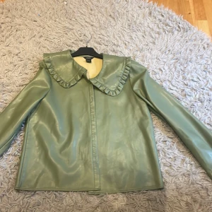 Grön jacka med volangkrage - Säljer en superfin grön jacka med en unik volangkrage. Jackan är långärmad och har en stilren design som passar perfekt för både vår och höst. Materialet ser ut att vara syntetiskt och ger en snygg glansig finish. Perfekt för dig som vill sticka ut lite extra!