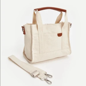 Beige axelväska i bomull - Säljer en superfin beige axelväska i bomull. Den har ett avtagbart axelband och handtag i brunt skinn. Perfekt för vardagsbruk med en stilren och enkel design. Väskan har en praktisk ficka på framsidan och stängs med dragkedja.