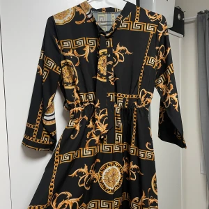 Svart och guldmönstrad långklänning - Säljer en elegant svart långklänning med ett iögonfallande mönster i guld. Klänningen har trekvartsärmar och ett knytband i halsen. Perfekt för en festlig kväll eller en speciell tillställning. Materialet känns mjukt och bekvämt mot huden.
