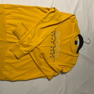 Gul tröja från Hugo Boss - Säljer en snygg gul tröja från Hugo Boss med deras logga tryckt på framsidan. Tröjan har långa ärmar och ribbade muddar vid ärmslut och nederkant. Perfekt för en färgglad stil!