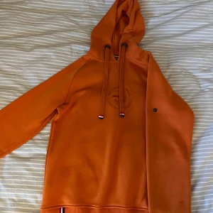 Orange hoodie från WHO's A.C.E. - Säljer en cool orange hoodie från WHO's A.C.E. med broderad logga på bröstet. Den har en justerbar huva med snören och en liten knappdetalj på ärmen. Inte använd någonting bara testad så inga tecken på användning. Nypris är 990
