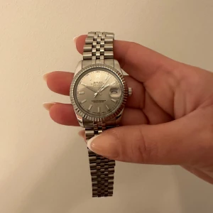 Silverfärgad klocka med länkarmband - Elegant silverfärgad klocka som är automatisk med ett klassiskt länkarmband. Urtavlan har en stilren design med datumvisning. Perfekt för den som söker en tidlös accessoar.