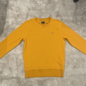 Gul tröja från GANT - Säljer en snygg gul tröja från GANT med rund hals och diskret logga på bröstet. Perfekt för en avslappnad stil. Tröjan har långa ärmar och ribbade muddar.