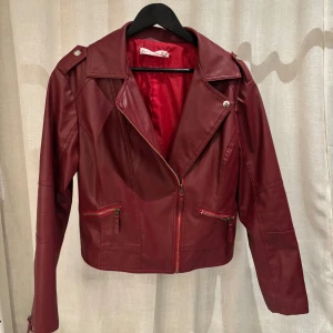 Vinröd skinnjacka med dragkedja - Snygg vinröd skinnjacka med dragkedja och långa ärmar. Jackan har en klassisk bikerstil med detaljer som axelklaffar och snedställd dragkedja. Perfekt för en cool och stilren look. Storleken är XXL men extremt liten k storlek och passar mer en S/M