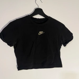 Svart croppad t-shirt från Nike - Snygg svart croppad t-shirt från Nike med korta ärmar och det klassiska Nike-loggan i vitt på bröstet. Perfekt för en sportig look eller avslappnad stil.