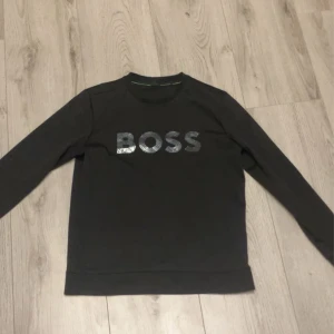 Svart tröja från Boss - Snygg svart tröja från Boss med glittrig logga på bröstet. Perfekt för en stilren look. Tröjan har långa ärmar och en klassisk passform.