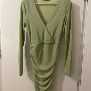 Glittrig grön omlottklänning - Snygg glittrig grön omlottklänning med v-ringning och långa ärmar. Perfekt för en kväll ute eller en speciell tillställning.