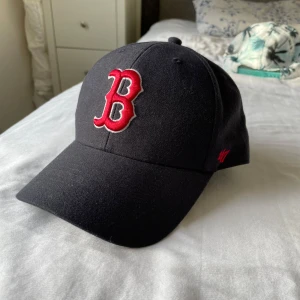 Svart keps med röd broderad B - Snygg svart keps från 47 med en stor röd broderad bokstav B på framsidan. Kepsen har justerbar rem baktill med texten 'Red Sox'. Perfekt för sportiga tillfällen eller som en stilren accessoar.