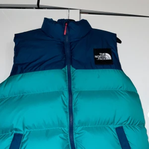 Blå och turkos dunväst från The North Face - Säljer en snygg blå och turkos dunväst från The North Face. Västen har en dragkedja framtill och är perfekt för kyligare väder. Den har en hög krage och märkets logga på bröstet.