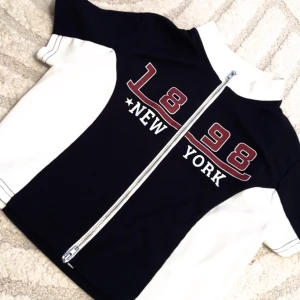 Svart och vit jacka med New York-tryck - Snygg svart och vit jacka med dragkedja och tryck '1898 New York' på framsidan. Perfekt för en sportig look.