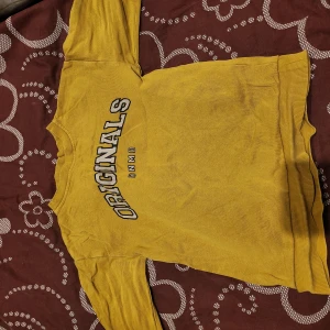 Gul tröja med tryck från Adidas - Säljer en gul tröja från Adidas med texten 'ORIGINALS DNMD' tryckt på framsidan. Tröjan har långa ärmar och ribbade muddar vid ärmslut och nederkant. Perfekt för en avslappnad stil.