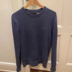 Blå tröja från Tommy Hilfiger - Säljer en stilren blå tröja från Tommy Hilfiger i Pima bomull och kashmir. Tröjan har en liten logotyp på bröstet och ribbade kanter vid hals, ärmslut och nederkant. Perfekt för en avslappnad men snygg look.