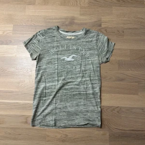 Gråmelerad t-shirt från Hollister - Snygg gråmelerad t-shirt från Hollister med tryck av deras ikoniska logga och texten 'California'. Perfekt för en avslappnad stil. Kortärmad och tillverkad i mjukt material för extra komfort.