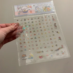 Klistermärken med Sumikko Gurashi-motiv - Ett ark med söta klistermärken från San-X med Sumikko Gurashi-karaktärer. Klistermärkena har holografiska stjärnor som ger en glittrande effekt. Perfekt för att dekorera anteckningsböcker, brev eller andra ytor.