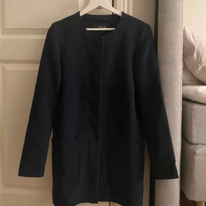 Mörkblå kappa från Vila - Elegant mörkblå kappa från Vila med en stilren design. Kappan har långa ärmar och en rak passform, perfekt för höst och vår. Den är tillverkad i ett mjukt material och har diskreta fickor framtill.