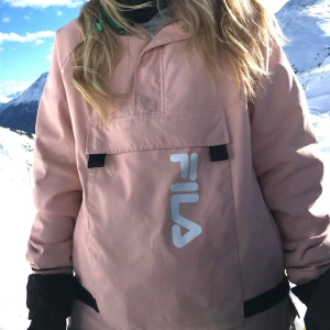 Rosa vindjacka från FILA - Snygg rosa vindjacka från FILA med stor ficka framtill och justerbar huva. Perfekt för skidåkning eller blåsiga dagar och har en sportig look med logga på bröstet.