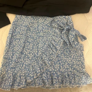 Blå blommig omlottkjol -  blå omlottkjol med ett sött blommönster. Kjolen har volangdetaljer och ett knytband i midjan för en justerbar passform. Använd ca 2 gånger och säljer då jag inte vill ha den mera.