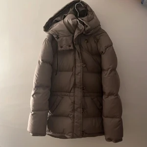 Brun dunjacka från Boomerang - Säljer en klassisk brun dunjacka från Boomerang i storlek XS. Jackan har en huva och stängs med både dragkedja och knappar. Perfekt för kalla dagar med sin varma dunstoppning. Ordinarie pris 5000kr.