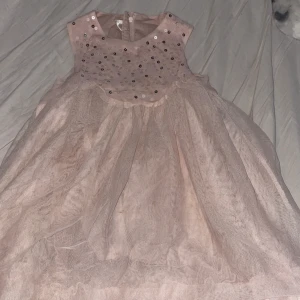  Barn  Rosa klänning med paljetter - Charmig rosa klänning med glittriga paljetter på överdelen. Den har en luftig kjol i flera lager och är ärmlös, vilket ger en söt och festlig look. Perfekt för speciella tillfällen.