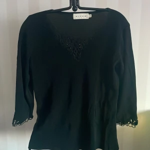 Svart topp från MICHA st. M - Elegant svart topp från MICHA med v-ringad spetsdetalj och trekvartsärmar i spetsdetalj. På etiketten står det 44 den är mer en M.Perfekt för en stilren look med fina detaljer vid ärmslut.