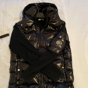 Svart dunväst från Moncler - Snygg svart dunväst från Moncler med glansig yta och dragkedja framtill. Västen har en hög krage och tryckknappar på fickorna. Perfekt för kyliga dagar när du vill hålla stilen.