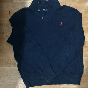 Mörkblå tröja från Ralph Lauren - Säljer en stilren mörkblå tröja från Ralph Lauren med knappar vid halsen och det klassiska röda logotypbroderiet på bröstet. Tröjan har långa ärmar och ribbade muddar för en bekväm passform.